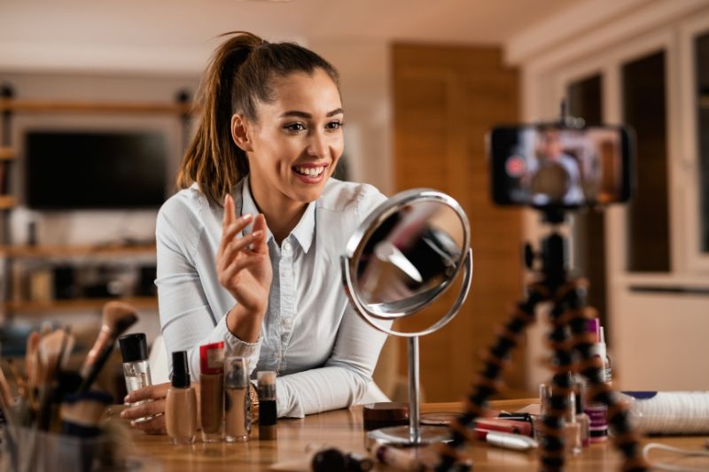 Mujer influencer sonriendo mientras graba una sesión de maquillaje con el celular en un tripié